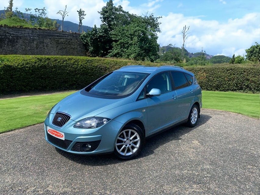 A blue compact hatchback car parked on a gravel drive with green manicured lawns and hedges in the background under a clear sky.