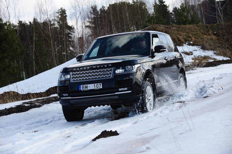 A Range Rover driving through a snow-laden path, splattering snow as it moves. The surroundings are forested and the sky is overcast. An individual is noticeable driving the vehicle.
