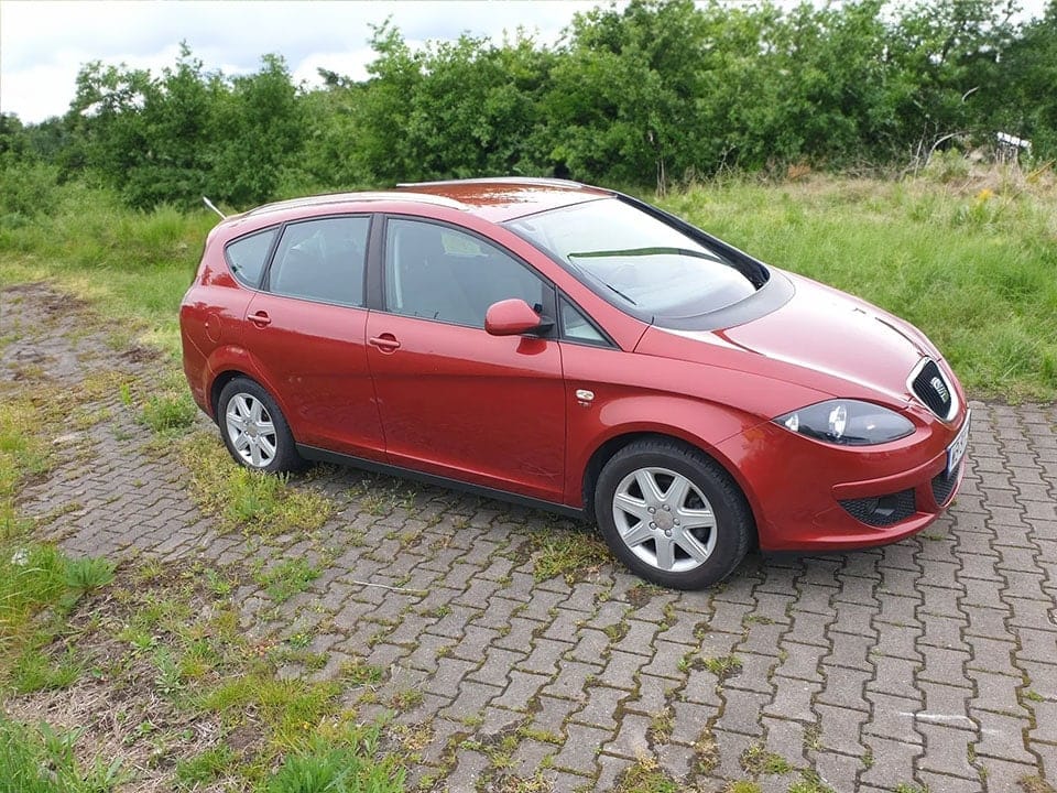 A red hatchback car parked on a patchy grass area with a paved stone path, surrounded by greenery under a cloudy sky.