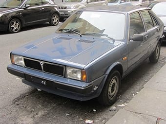 A blue saloon parked on a city street. The car is an older model with a noticeable bonnet vent and European number plates.