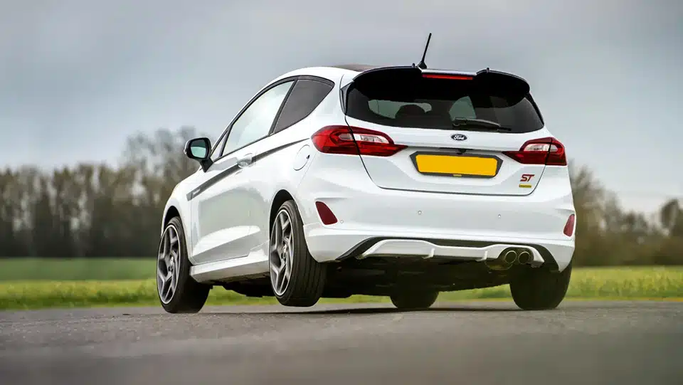 A white Ford Fiesta ST parked on an open road with a grassy field in the background, showcasing its rear design and dual exhaust. Fitted with Polybush Rear Axle Bushes