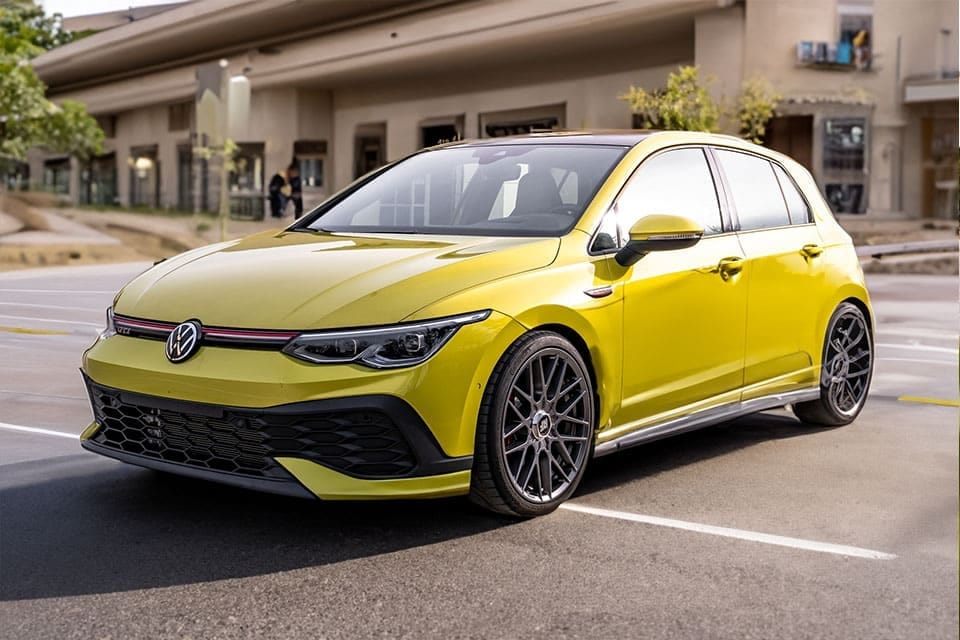 A bright yellow Volkswagen Golf GTI parked on a road, boasting sleek lines and sporty black alloys, captured on a cloudy day with a gently blurred background highlighting buildings.