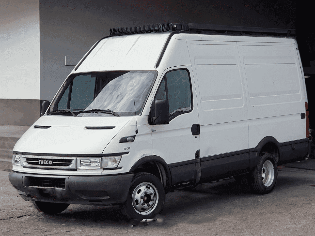 A white Iveco Daily van parked on a concrete surface near a building, featuring a sliding side door and roof rails.