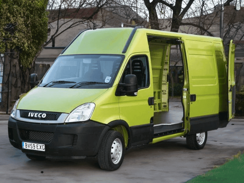 A vibrant lime green Iveco van parked with its sliding door open, offering a peek of the interior, positioned against a residential backdrop with leafless trees and a hedge.