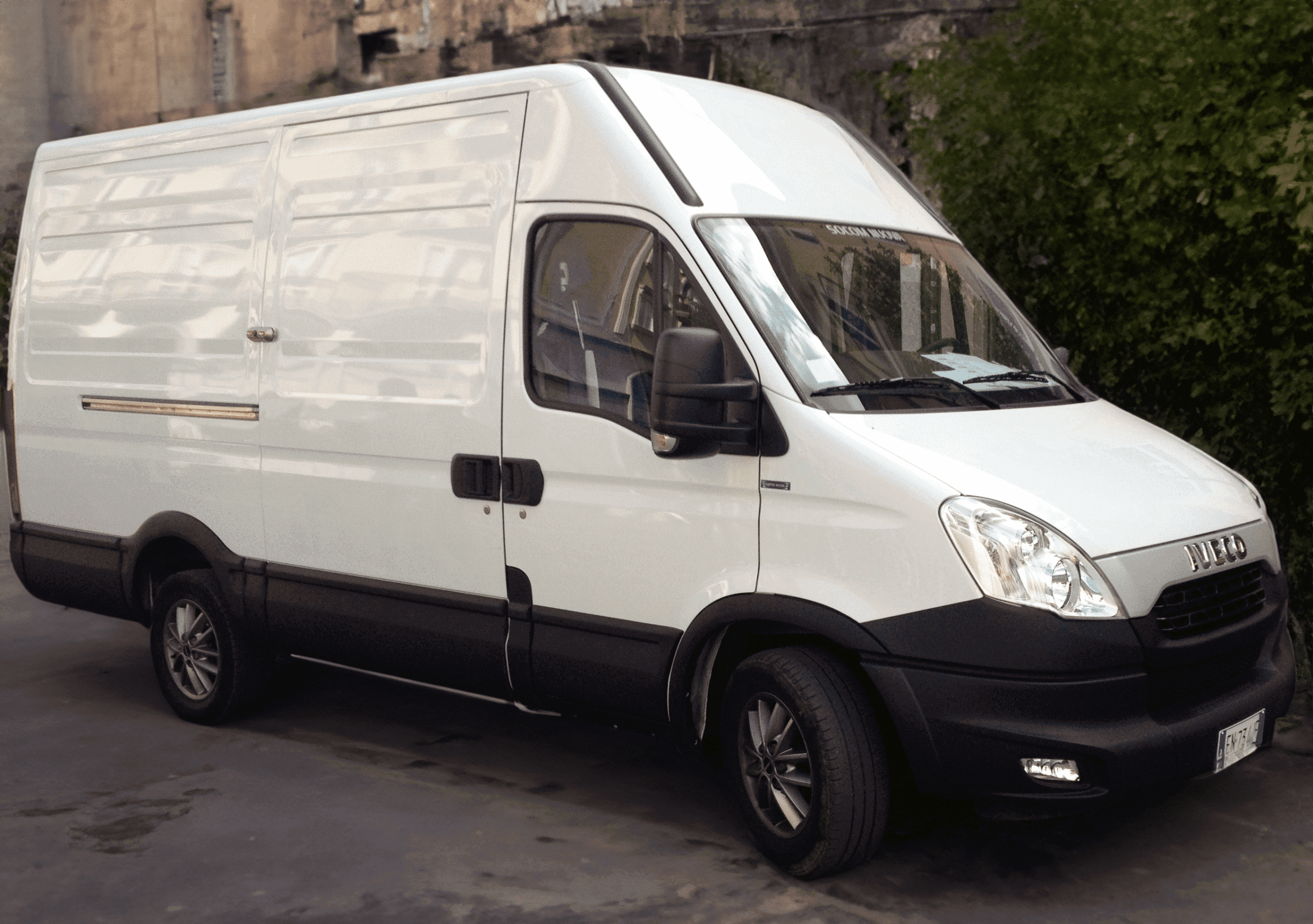 A white delivery van parked on a concrete surface, featuring a sliding door and pronounced black wheels, with shadow reflections on its side.