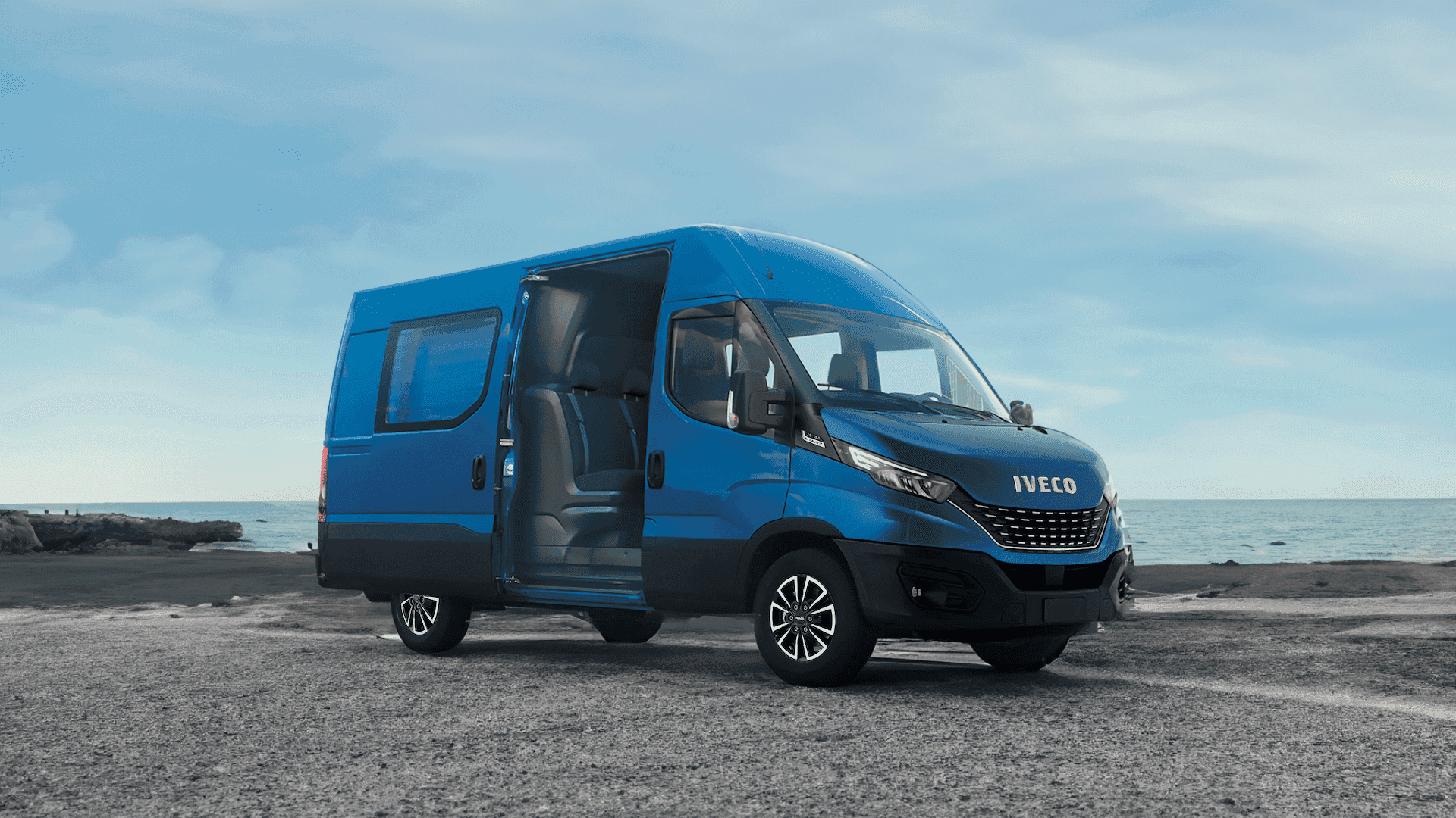 A blue Iveco van parked by the seaside with the driver's door open, showing the interior. The backdrop features a clear sky and rocky coastline.
