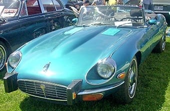 A classic teal Jaguar E-Type convertible parked at a car show, showcasing its sleek design with chrome bumpers and wire wheels under bright sunlight.