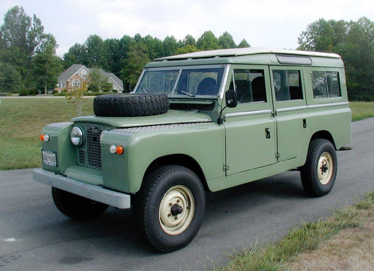 A classic green Land Rover Series II parked on a kerb with grass and houses in the background. The vehicle features a spare tyre mounted on the front bonnet.