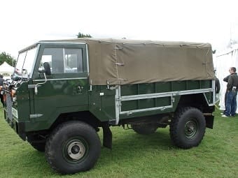 A green military lorry with a canvas-covered cargo area parked on grass at an outdoor event. The lorry has large off-road tyres and a basic boxy design.