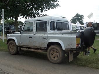 A muddy white Land Rover Defender with four doors parked on a grassy area at an outdoor event, with a partial view of a man walking beside it.