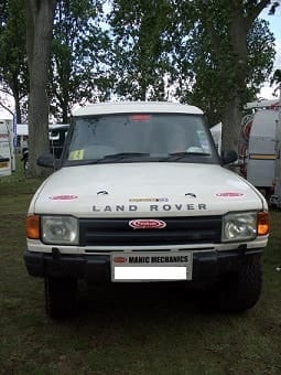 Front view of a white Land Rover Discovery with sponsorship stickers, parked on grass at an outdoor event. Visible trees and other vehicles are in the backdrop.