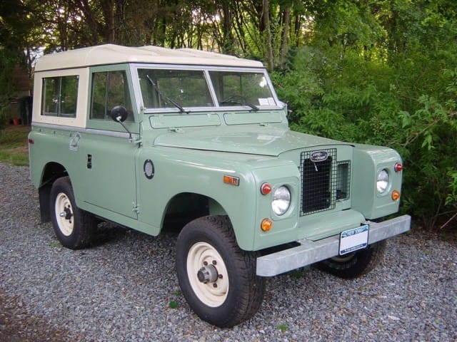 A vintage green Land Rover Series IIA parked on a gravel drive, surrounded by trees, featuring a white roof and tyres with cream-coloured hubs.
