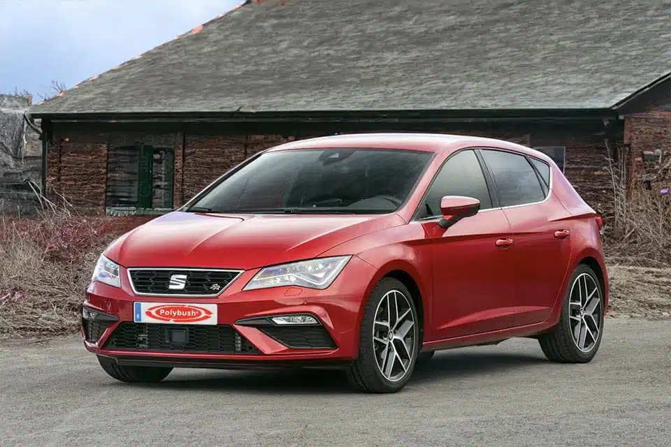 A bright red Seat Leon parked in front of an old brick building with a dark, slanted roof. The car is modern and sleek, featuring sharp lines and sporty design elements.