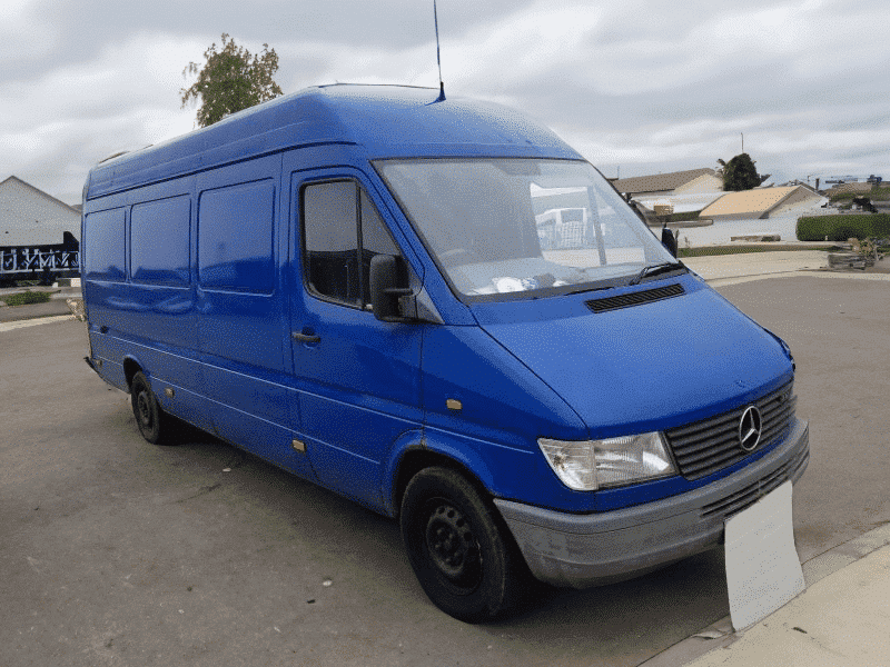 A blue Mercedes-Benz Sprinter van parked on a tarmac surface with overcast skies in the background. The van features a high roof and tinted windows.