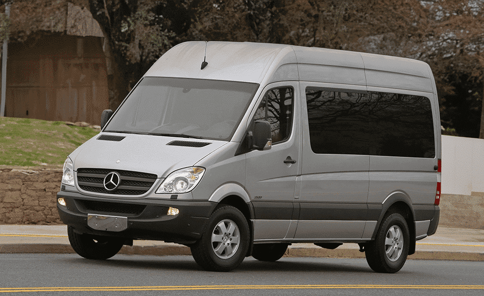 A silver Mercedes-Benz Sprinter van parked on a roadside with trees in the background. The vehicle is modern and features tinted windows.