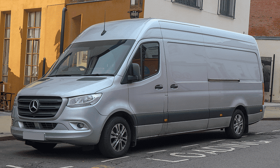 A silver Mercedes-Benz Sprinter van parked on a road, featuring a high roof and sliding side door, with a bright yellow building in the backdrop.