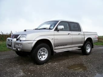 A silver Mitsubishi L200 Warrior pickup lorry parked on a gravel road with a cloudy sky in the background.