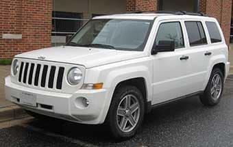 A white jeep patriot parked on a wet road next to a brick building, on an overcast day.