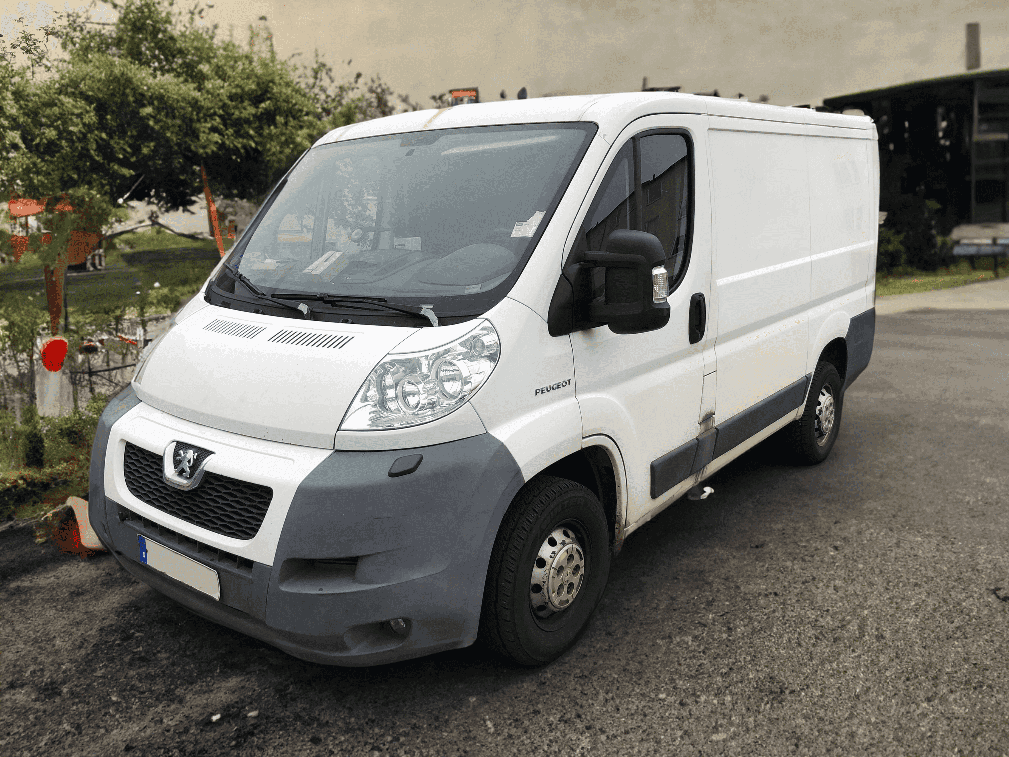 A white Peugeot Boxer delivery van parked on a paved area near a workshop, featuring a partially grey front bumper and visible signs of wear and tear.