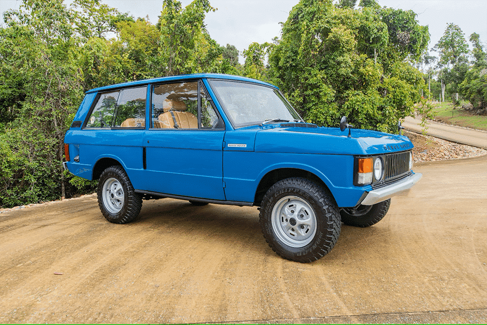 A vintage blue range rover parked on a concrete road surrounded by lush vegetation. The vehicle looks well-maintained, showcasing classic design and features.