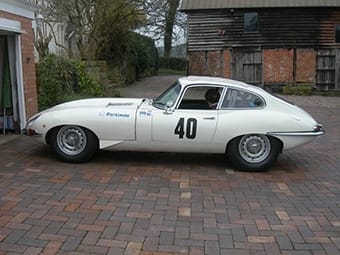 A vintage white Jaguar E-type coupe with the number 40 on its side, parked on a brick drive beside wooden barn structures, with a person visible inside the car.