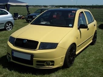 A yellow Skoda Fabia parked on a grass field under clear skies, featuring tinted windows and black alloy wheels. Other cars and tents in the background suggest a motor show setting.