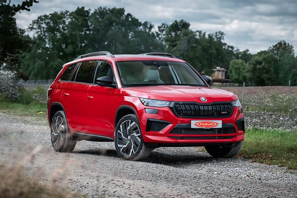 A red SUV parked on a gravel track in a rural area, showcasing a modern design with sharp lines and distinctive lighting. Verdant green trees and a cloudy sky can be seen in the background.
