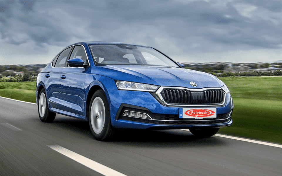 A blue Skoda saloon is driving on a road, with lush green grass and trees blurred by motion in the backdrop, under a cloudy sky.