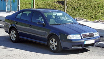 A blue saloon car parked on the side of a road during daylight, with a green grassy area and a metal barrier in the background. The car features a clean design with visible headlamps and a grille.