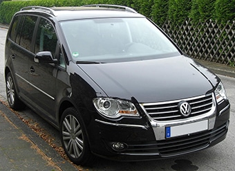 A black Volkswagen Touran MPV parked on a wet street, featuring a prominent chrome grille and dual headlights, with hedges in the background.