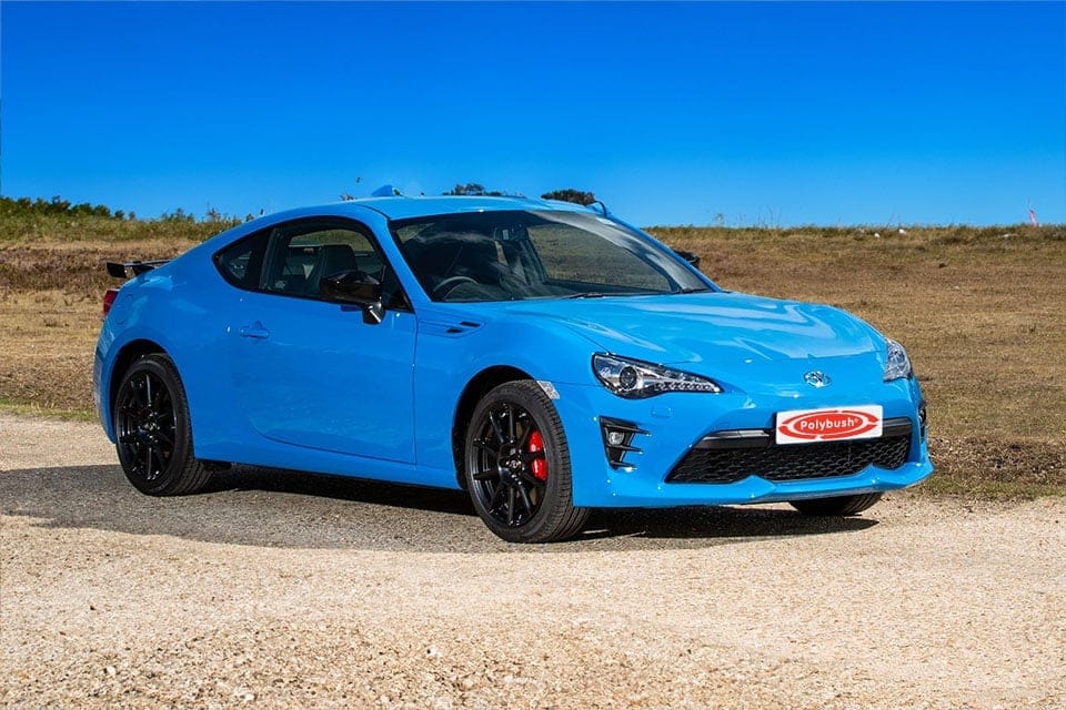 A bright blue sports car, possibly a Subaru BRZ, parked on a gravelly surface with a clear blue sky and sparse vegetation in the background. The car features black alloys and visible branding.
