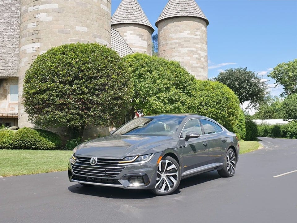 A sleek grey Volkswagen Arteon parked on a road, with lush green trees and two round stone towers in the background under a clear blue sky.