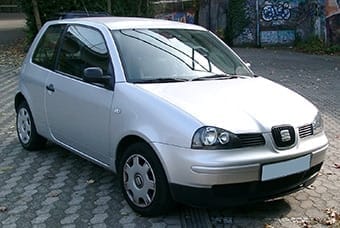 A compact silver hatchback car parked on a cobbled street, featuring a distinctive round logo on the grille and framed by graffiti-covered walls.