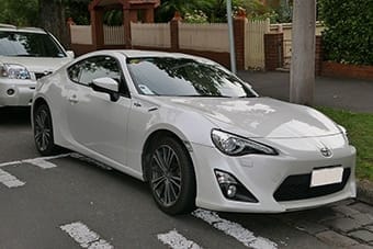 A white sports coupé parked on a suburban street, with other cars and greenery in the background.