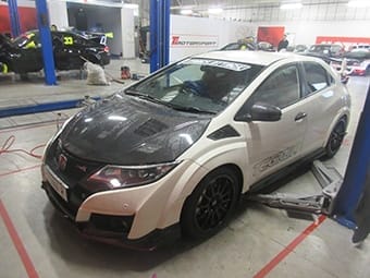 A Honda Civic with a black carbon fibre bonnet and white body, featuring racing decals, parked inside a busy garage with various cars and equipment in the background.