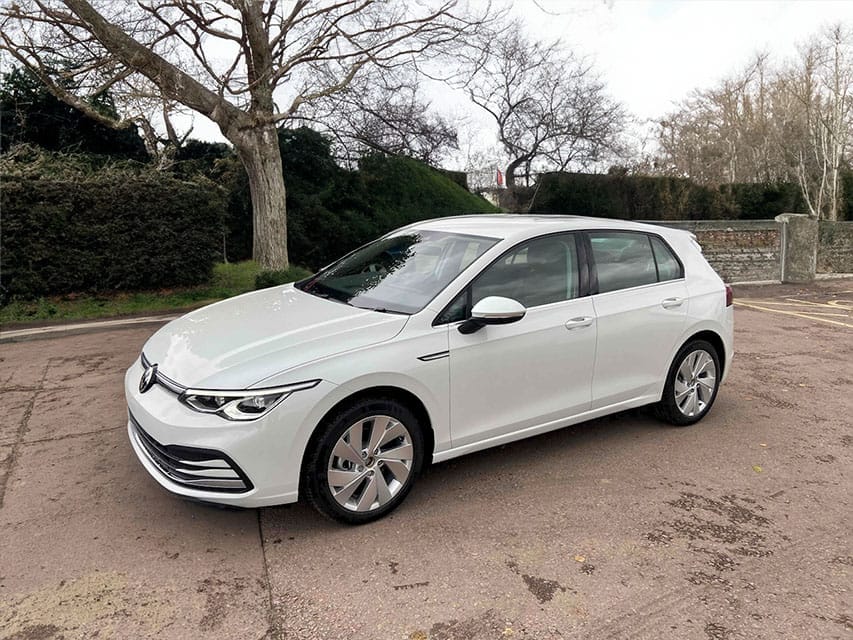 A white contemporary electric hatchback car parked on a gravelled area with trees and a fence in the background on a cloudy day.
