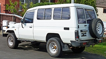 A white, four-door SUV parked on a residential road, with a spare tyre mounted on the back. The vehicle has apparent off-road capabilities and is situated under a bright, clear sky.