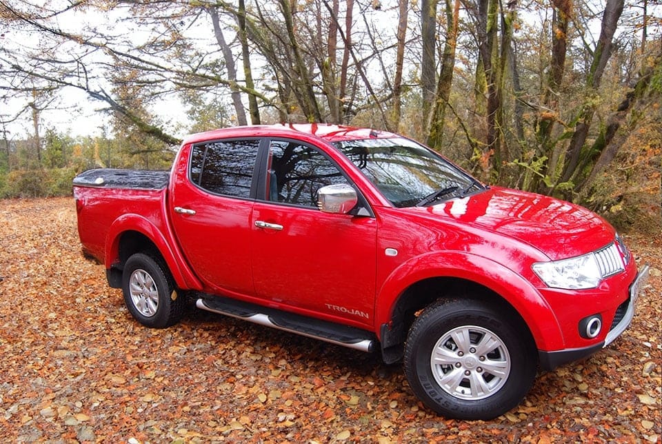 A red pick-up lorry parked on a gravel path surrounded by autumnal trees with fallen leaves.