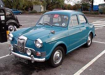 A classic teal blue Riley One-Point-Five saloon parked on a street, with a vintage black taxi visible in the background.