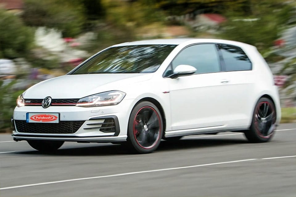 A white Volkswagen Golf GTI moving swiftly on a blurred road, exhibiting its sporty design and dynamic performance. The car features distinctive red trim on wheels and a black roof.