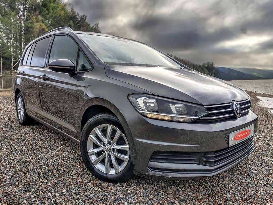 A dark grey Volkswagen Golf parked on a gravelled car park, with cloudy skies and a forested mountain in the background.