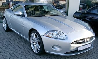 A silver sports coupé parked outside, featuring sleek lines, large headlamps, and alloy wheels, with a clear blue sky reflected on its polished surface.