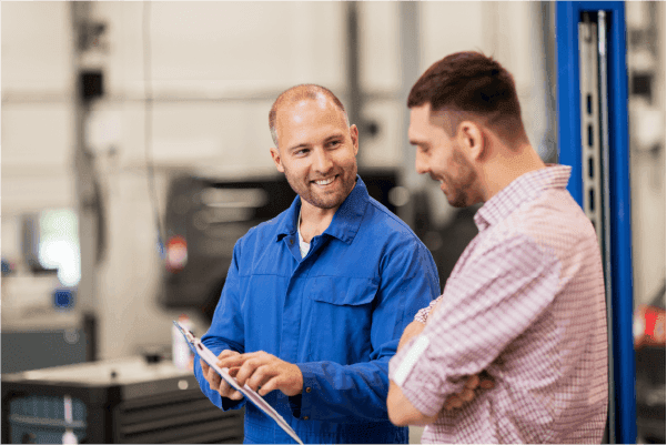 Two men in a workshop discussing over a clipboard; one in a blue mechanic overall and the other in a casual shirt, both smiling and engaged in conversation.