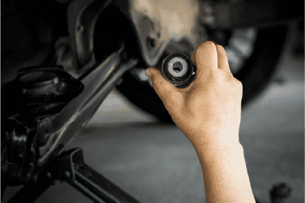 A person holding a car part near the wheel assembly of a vehicle in a garage, focusing on motor maintenance or repair.