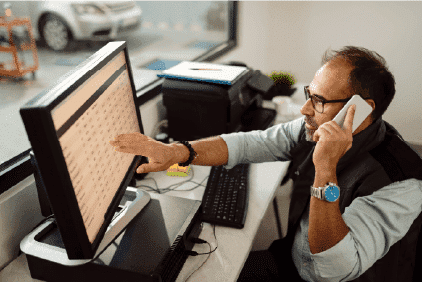 A middle-aged man wearing spectacles and using a phone while pointing at a computer screen in an office environment.