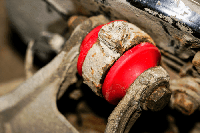 Close-up image of a red, worn-out rubber bushing on a vehicle's suspension system, showing visible signs of corrosion and dirt build-up.