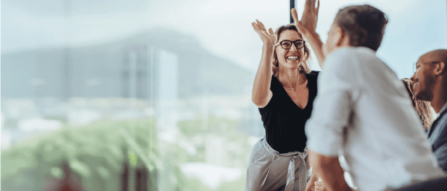 A joyful woman with spectacles high-fiving a colleague in a contemporary office setting with large windows and a blurred cityscape in the background.