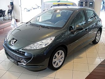 A Peugeot 207 displayed inside a showroom. The car is metallic grey, featuring a prominent logo on the grille, sleek headlights, and a streamlined design.