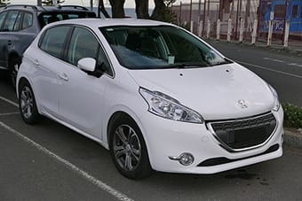 A white Peugeot 208 parked on a kerbside, featuring a sleek design with alloy wheels and visible headlamps, situated between a black vehicle and a construction barrier.