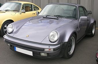 A metallic grey Porsche 911, an iconic sports car, parked between two other vehicles, showcasing its distinctive round headlights and smooth, sleek lines.